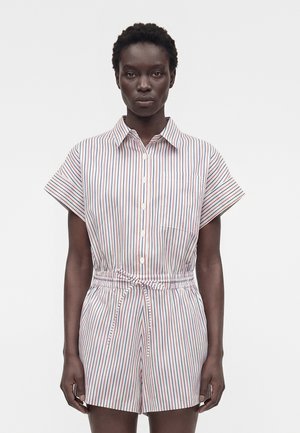 Nő fehér rövid ujjú, gombos kezeslábasban, piros és kék függőleges csíkokkal, rugalmas derékkal és elöl megkötve, fehér háttér előtt áll.