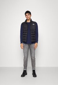 Svart polstret vest over en marineblå langermet skjorte, grå jeans og svarte snørebåndsstøvler. Modellen står mot en ensfarget bakgrunn.