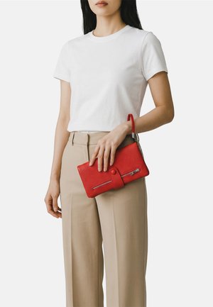 Donna che indossa una maglietta bianca e pantaloni beige che tiene una pochette rossa testurizzata con cerniera e cinturino da polso.