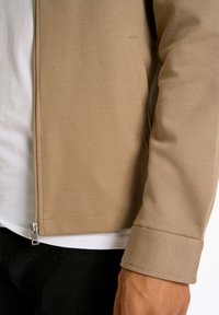 Chaqueta beige con cremallera hecha de tejido texturizado, que presenta un cuello alto, bolsillos laterales y una camiseta blanca visible debajo.