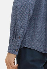 Blaues Langarmhemd mit geometrischem Druck in Rot und Weiß, mit geknöpften Manschetten und abgerundetem Saum, aus leichtem Stoff.