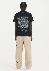 Czarna koszulka z dużym srebrnym graficznym wizerunkiem tygrysa na plecach, w połączeniu z luźnymi beżowymi spodniami zdobionymi kolorowymi haftami i akcentami z logo.