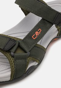 Green textile sandal featuring adjustable straps, texture contrast, gray cushioned footbed, and a black rubber sole with orange accents.