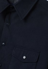 Primer plano de un abrigo de lana azul marino oscuro con cuello afilado, botones a presión y un bolsillo de solapa que cuenta con un único botón a presión.