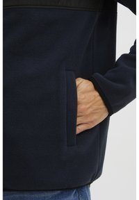 Hand rustend in de zak van een marineblauwe fleecejas, gedragen met een blauwe spijkerbroek, op een lichte achtergrond.