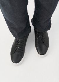 Μαύρα δερμάτινα αθλητικά παπούτσια με υφή, στρογγυλή μύτη και λευκή καουτσούκ σόλα, βλέπουμε από πάνω, συνδυασμένα με σκούρα τζιν παντελόνια.