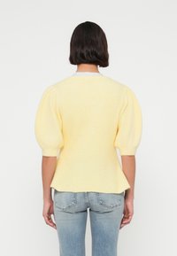 Κίτρινο πλεκτό πουλόβερ με φουσκωτά κοντά μανίκια, υφασμάτινη υφή και εφαρμοστή μέση. Συνδυάζεται με ανοιχτόχρωμα τζιν παντελόνια.