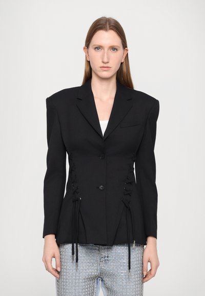 Schwarzer taillierter Blazer mit breiten Schultern, Knopfverschluss auf der Vorderseite und Schnürdetails an den Seiten, kombiniert mit hellen Jeans.