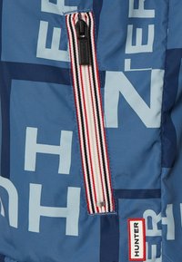 Niebieska pikowana kurtka z zamkniętą kieszenią z paskowanym akcentem i dużymi literami. Na dolnym końcu widoczna jest metka z logo w kolorze czerwonym.