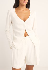 Blusa de lino blanca con escote en V profundo y cierre de botones, combinada con pantalones cortos blancos a juego. Textura suave y corte relajado.