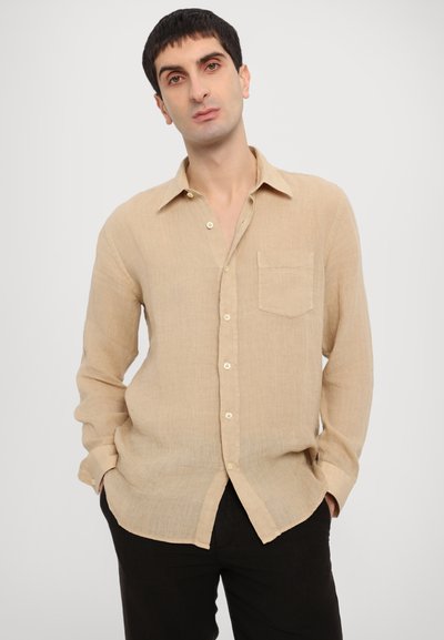 Camicia di lino beige con colletto button-down, maniche lunghe, taschino sul petto e vestibilità morbida, abbinata a pantaloni scuri.