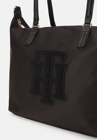 Black nylon tote bag featuring a prominent embroidered logo, leather straps, and gold hardware accents. Smooth texture with a structured shape.