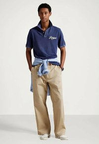 Młody mężczyzna stoi z rękami w kieszeniach, ubrany w granatową koszulkę polo z napisem "Ralph", beżowe spodnie, jasnoniebieski sweter przewiązany w talii i białe buty.