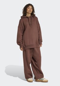 Brun oversized hoodie med framficka och dragsnöre, tillsammans med lösa bruna sweatpants. Tyget verkar mjukt och texturerat.
