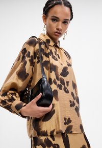 Bruine blouse met zwart dierenprint, knopenlijst en lange poofy mouwen. Een kleine zwarte handtas wordt vastgehouden met een schouderband.