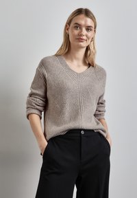 Beige gerippter Pullover mit V-Ausschnitt, mit umgeschlagenen Ärmeln und einem strukturierten Muster. Kombiniert mit schwarzen Hosen, die eine lässige Passform zeigen.