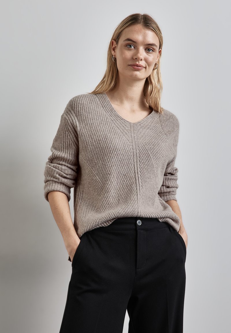 Beige gerippter Pullover mit V-Ausschnitt, mit umgeschlagenen Ärmeln und einem strukturierten Muster. Kombiniert mit schwarzen Hosen, die eine lässige Passform zeigen.