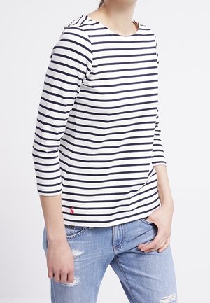 T-shirt à rayures bleu marine et blanches à manches longues avec un décolleté en forme de U et une texture côtelée ; présente un petit logo sur l'ourlet. Porté avec un jean délavé.