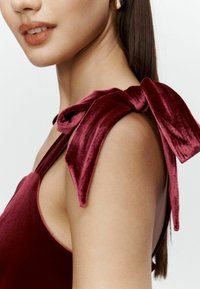 Burgundowa aksamitna odzież z dużą kokardą na ramieniu. Gładka tekstura; elegancka sylwetka podkreślająca ramię i dekolt.