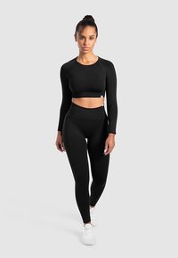 Svart crop top med långa ärmar och högmidjade leggings i slät, stretchig tyg. Modellen bär vita sneakers och står mot en neutral bakgrund.