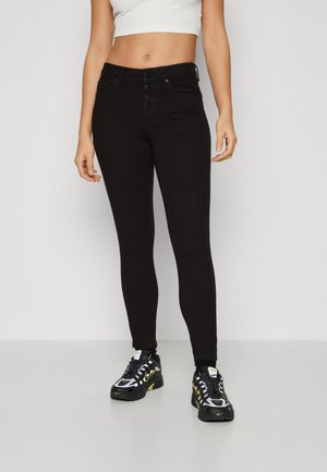 Black skinny jeans with a high waist, featuring five-button closure and front pockets. Worn with black and yellow athletic shoes.