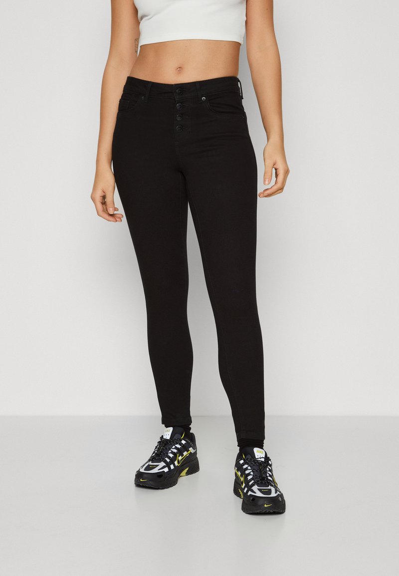 Jeans skinny negros de cintura alta, con cierre de cinco botones y bolsillos frontales. Combinados con zapatillas deportivas negras y amarillas.