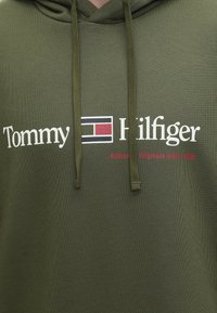 Λαδί φούτερ με κουκούλα με λογότυπο Tommy Hilfiger σε λευκό και κόκκινο κείμενο "Authentic Originals since 1985" μπροστά, συμπεριλαμβανομένου του συμβόλου σημαίας της μάρκας.