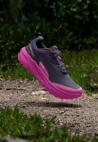 Μαύρο και ροζ παπούτσι για τρέξιμο σε μονοπάτι στον αέρα πάνω από χαλίκι, με φόντο πράσινη βλάστηση, που δείχνει την υφή της σόλας και τα κορδόνια.