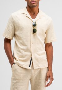 Beige kortemouwen overhemd met knopen, gemaakt van textuurstof, met een camp-kraag en een label aan de zijkant. Zonnebril ligt op de kraag.