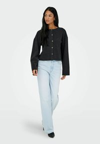 Cardigan nero corto con maniche lunghe e bottoni in legno, abbinato a jeans blu chiaro a gamba dritta e scarpe nere. Texture piatta, stile casual.