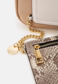 Chaîne dorée reliant un sac beige et blanc à une petite pochette à motif peau de serpent avec une tirette de fermeture éclair dorée et un charm ALDO.