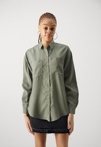 Olive green button-up shirt with long sleeves, two chest pockets adorned with silver studs, and a curved hem. Paired with a black denim skirt.