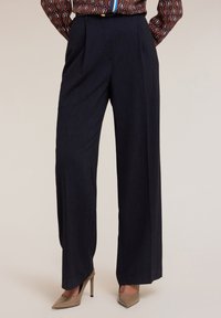 Tmavě modré kalhoty širokého střihu s vysokým pasem a předními záhyby, vyrobené z texturované látky, spárované s béžovými vysokými podpatky.