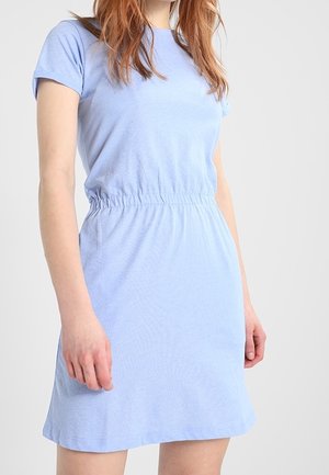 Femme porte une robe bleu clair à manches courtes avec une taille élastique, debout devant un fond clair uni.
