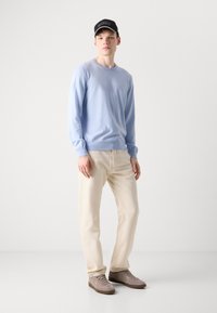 Sweater de crewneck azul claro, calças bege e sapatilhas de camurça cinza. O modelo usa um boné preto com detalhes de logo branco.