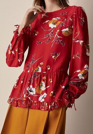 Kvinde iført en langærmet rød bluse med blomsterprint, flæsekant og bundne manchetter, kombineret med bukser i sennepsfarve.