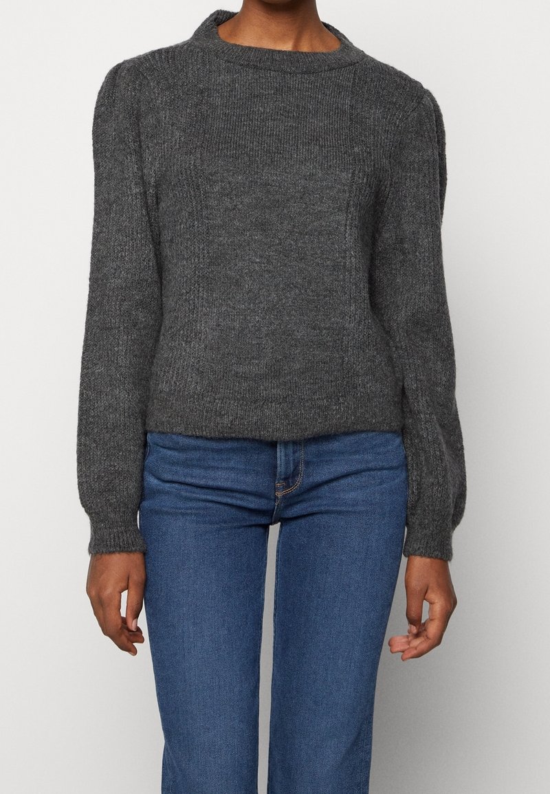 Personne portant un pull en tricot gris foncé à manches longues et un jean en denim bleu, debout, les bras détendus le long du corps.