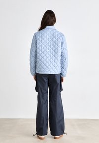 Personne aux cheveux longs portant une veste matelassée bleu clair et un pantalon cargo ample de couleur foncée, debout pieds nus sur un sol uni devant un mur blanc.