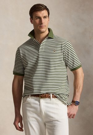 Mann iført grønn og hvit stripete poloshirt med hvite bukser og brunt flettet belte, stående med den ene hånden i lommen.