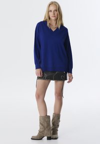 Königsblauer gestrickter Pullover mit V-Ausschnitt, kombiniert mit einem schwarzen Denim-Minirock mit Steppdetails und beigefarbenen, lässigen Knöchelstiefeln.