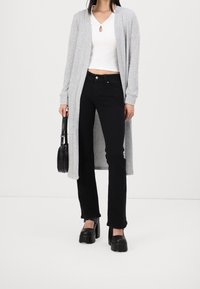 Kobieta ubrana w czarne spodnie z wysokim stanem, biały top do pępka, długi szary kardigan, czarne buty na platformie, trzymająca małą czarną torebkę.