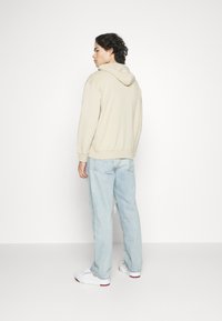 Beige hette med snörning, ribbede mansjetter og søm, kombinert med lyseblå slitte jeans og hvite joggesko med røde detaljer.
