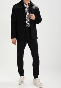 Černá bunda s koženými rameny, design na knoflíky; vzorovaná košile pod ní; černé tepláky; bílé sneakers. Minimalistický a přizpůsobený vzhled.