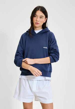 Młoda kobieta poprawiająca rękaw, ubrana w granatową bluzę z kapturem ellesse, białą koszulę pod spodem oraz białe szorty na jednolitym tle.