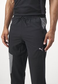 Zwarte sportbroek met een elastische tailleband, grijze zijkanten, ritszakken en een klein roze Puma-logo op de dij.