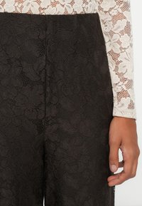 Pantalon en dentelle noire porté avec un haut en dentelle blanche à manches longues à motif floral, montrant des textures de tissu détaillées et une main détendue reposant sur le côté.