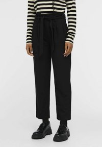 Pantalon noir ajusté avec une ceinture nouée à la taille, présentant des plis à l'avant et une coupe décontractée. Associé à des chaussures à plateforme noires et un haut rayé.