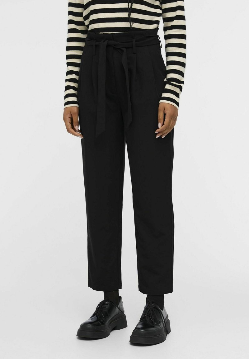 Pantalon noir ajusté avec une ceinture nouée à la taille, présentant des plis à l'avant et une coupe décontractée. Associé à des chaussures à plateforme noires et un haut rayé.