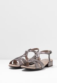 Sandales à talons bas argentées métalliques avec orteils ouverts, plusieurs lanières et fermetures à boucle au niveau de la cheville sur une surface blanche.