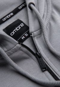 Sweat à capuche gris avec fermeture éclair noire et cordons, avec étiquette de marque « ombre » et étiquette taille M (UE 48) à l’intérieur du col.